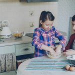 Little cheerful and funny girl helping her mother in the kitchen cropping egg into bowl and mixing dough for cookies. Family, food, home and people concept
